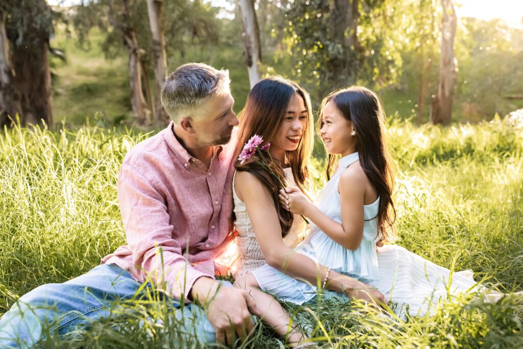 Biracial family of 3 sitting in tall grass laughing and smelling flowers with the sun setting behind them in Murrieta California
